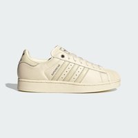 Ανοιχτόχρωνο αθλητικό παπούτσι Adidas Superstar με προστατευτική μύτη, τρεις ρίγες στο πλάι και το κείμενο "SUPERSTAR", εμφανιζόμενο σε πλευρικό προφίλ σε ανοιχτό φόντο.