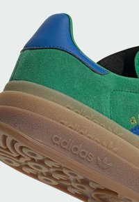 Sneaker verde em camurça com detalhes em couro azul, sola de borracha transparente e logótipo em relevo; apresenta uma forma arredondada e um upper texturizado.