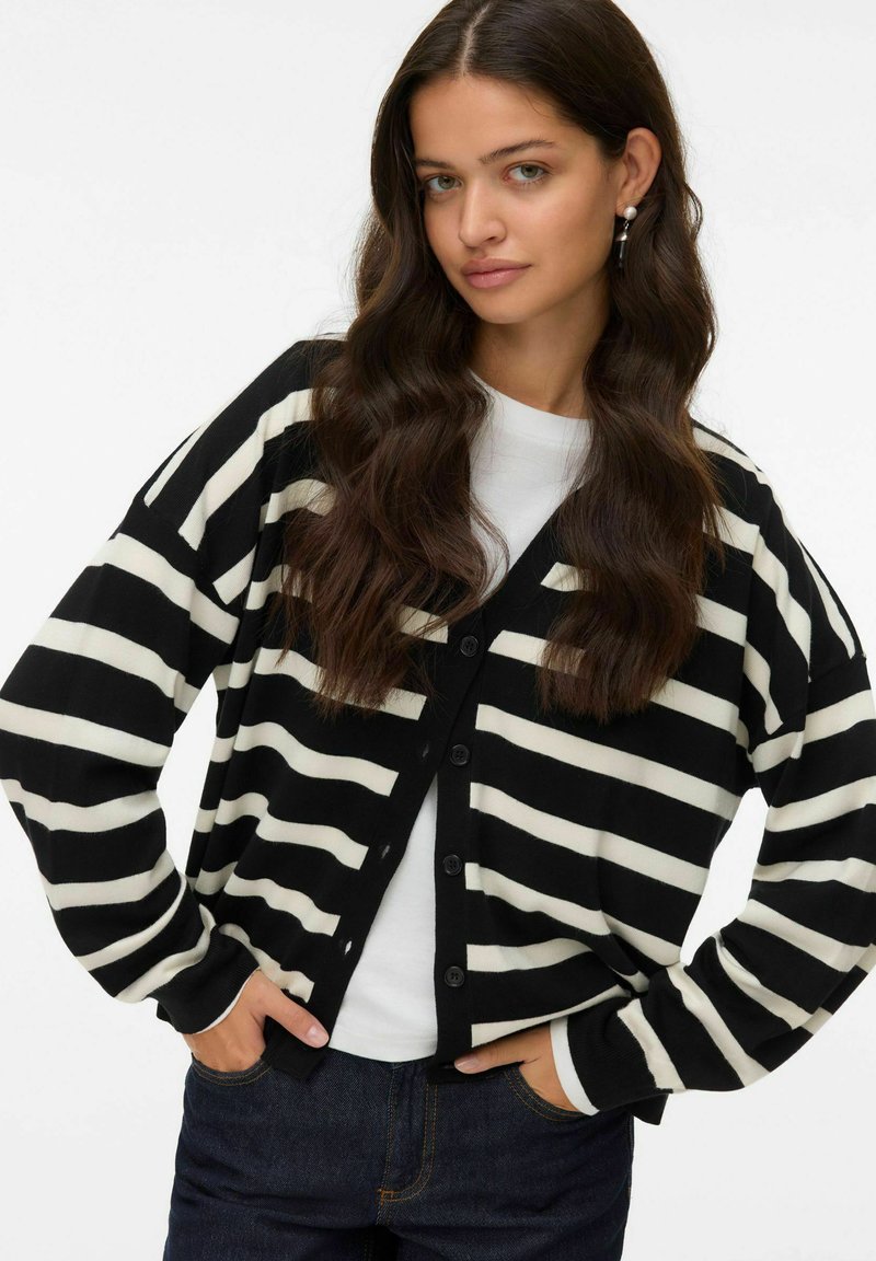 Cardigan rayé noir et blanc avec un col en V, des boutons sur le devant et des manches longues, porté sur une chemise blanche, associé à un jean foncé.