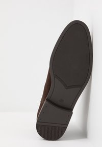 Zapato de ante marrón con una suela de goma texturizada. La suela tiene un acabado suave y un patrón distintivo cerca del talón para mejorar la tracción.