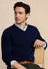 Granatowy wełniany sweter w serek z prążkowanymi mankietami, noszony na jasnoniebieskiej koszuli w paski. Model siedzi na drewnianym krześle z rattanowymi akcentami.