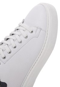 Zapatilla de cuero blanco con cordones blancos, ojales plateados, suela texturizada y costuras detalladas en el panel lateral.