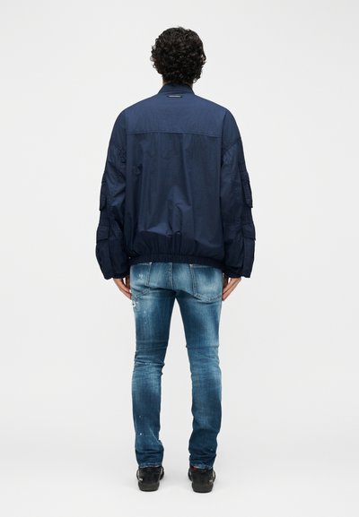 Άνδρας με σγουρά μαλλιά που στέκεται, φορώντας μπλε σκούρο μπουφάν με ελαστική μέση και ξεθωριασμένο μπλε τζιν, γυρισμένος πλάτη σε απλό φόντο.