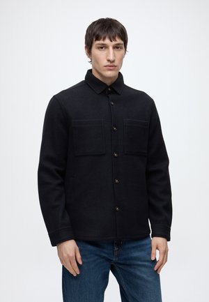Mann mit kurzen braunen Haaren, der eine schwarze Jacke mit Knöpfen und Brusttaschen sowie blaue Jeans trägt, steht vor einem weißen Hintergrund.