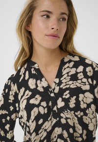 Zwarte blouse met een beige abstract patroon, v-hals en drie zwarte knopen aan de voorkant. Zachte textuur en ontspannen pasvorm.