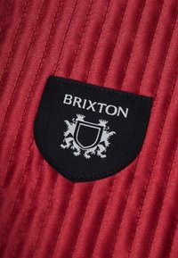 Piros szövet függőleges bordázattal, fekete pajzs folttal, rajta a "BRIXTON" felirattal egy részletes címer felett, fehér színben.