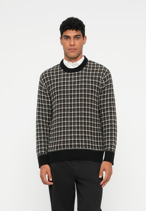 Uomo con un maglione a maglia a scacchi bianco e nero sopra una camicia bianca con colletto, abbinato a pantaloni neri, che si trova davanti a uno sfondo neutro.