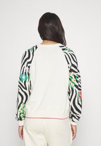 Pull mettant en avant un corps crème avec des manches imprimées zèbre en noir et blanc, ornées de motifs floraux rouges et bleus, avec des poignets et un ourlet côtelés.