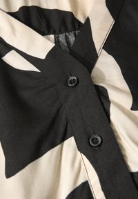 Camicia nera e beige con bottoni, caratterizzata da un motivo geometrico, in morbido tessuto, con scollo a V e pieghe attorno all'area dei bottoni.