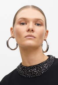 Svart topp med pärlkrage och stora silverfärgade hoopörhängen. Slät tygtextur och rund halsringning. Närbild av modell.