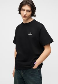 Νεαρός με μεσαίου μήκους μαλλιά που φοράει μαύρο μπλουζάκι με μικρό λευκό λογότυπο "JACKER" και σκούρο τζιν, στέκεται με ένα χέρι στην τσέπη.