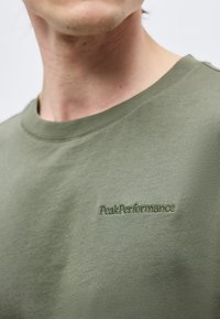 Gros plan sur une personne portant un t-shirt col rond vert olive avec « Peak Performance » brodé sur la poitrine.