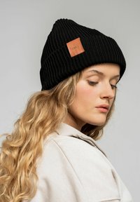 Czarna dzianinowa czapka z prążkowanym splotem, z brązową skórzaną łatką. Projekt charakteryzuje się dopasowanym kształtem i teksturowanym materiałem.