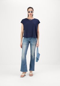 Marineblå kortermet topp, lyseblå flagrende jeans, lyse sandaler og en blå håndveske. Tilbehøret inkluderer runde hvite solbriller og et smykke.