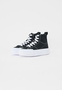 Svart høyhælte sneakers laget av lerret med hvite sømmer, hvite gummisåler og snørebånd. Har to løftestropper bak.
