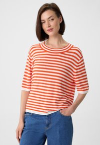 Pasiasta koszulka w kolorze pomarańczowym i białym, okrągły dekolt, krótkie rękawy, luźny krój, miękki materiał i kontrastowe ściągacze, w zestawie z niebieskimi dżinsami.