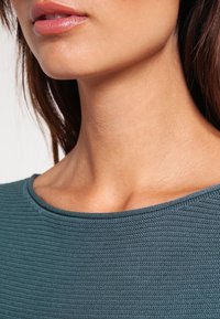 Turquoise geribbelde trui met een ronde halslijn, gestructureerde stof en een gladde afwerking. Close-up gericht op de halslijn en de kraag.