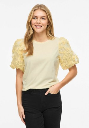 Femme aux longs cheveux blonds portant un haut jaune pâle aux manches bouffantes texturées et un pantalon noir, souriant avec une main dans la poche.