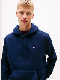 Young man with short blonde hair wearing a navy blue hoodie with a small red, white, and blue logo on the chest, standing against a plain background.