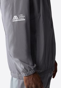 Harmaa kevyt takki, jossa on joustavat hihansuut ja helma, ja The North Face -logo hihassa, valmistettu sileästä synteettisestä materiaalista.