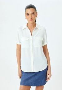 Witte korte mouwen shirt met twee voorzakken, knoopsluiting aan de voorkant en een gladde textuur. Gecombineerd met een blauwe gestructureerde rok.