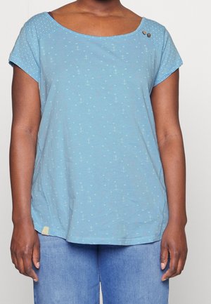 Mujer con una camiseta de manga corta azul clara con pequeños anclas y patrones de corazones y jeans azul claro, de pie con los brazos relajados.