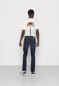 Kortärmad krämfärgad t-shirt med en färgstark grafisk design på ryggen, kombinerad med mörkblå jeans och vita sneakers.