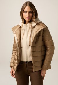 Puffer jacket in light brown with horizontal quilted pattern, featuring a detachable hood and gold-tone hardware. Worn over a cream hoodie.