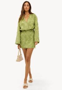 Tenue vert clair en tissu texturé, comprenant un haut croisé et une mini jupe. Accessoirisée avec un petit sac beige et des sandales plates.