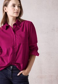Fuchsia button-up shirt with a pocket, rolled sleeves, and soft texture, paired with dark denim jeans featuring zippers and contrast stitching.