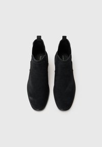 Zapatos negros de ante sin cordones con una textura suave, puntera redondeada, pequeña lengüeta y suela de goma plana. Diseño simple sin acentos visibles.