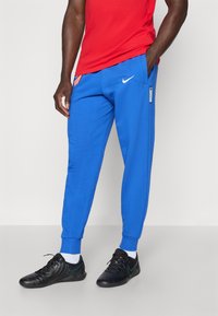 Blue athletic joggers made of soft fabric, featuring side pockets, a contrasting logo, and elastic cuffs. Paired with black athletic shoes.