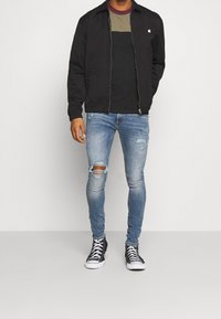 Svart zip-up jacka över en svart och olivfärgad blockfärgad t-shirt. Bärs med slitna blå skinny jeans och svarta högstövel sneakers.