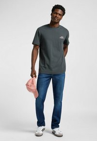 Czarna szara koszulka z nadrukowanym logo na klatce piersiowej, zestawiona z niebieskimi dżinsami i jasnoróżową czapką bejsbolową trzymaną w jednej ręce.