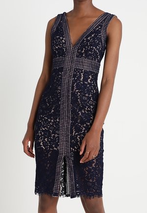 Robe sans manches en dentelle bleu marine avec un décolleté plongeant en V, une bordure à motifs au centre et à la taille, et une fente à l'avant, portée par une personne debout.