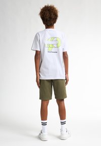 Tricou alb cu un grafic galben neon pe spate, asortat cu pantaloni scurți verzi măslinii, ciorapi cu dungi alb-negru și adidași albi.