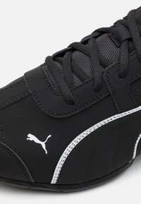 Musta urheilujalkine, jossa on strukturoidut yläosat, valkoiset tikkaukset ja pieni ikoninen logo. Pyöristetty kärki ja nauhakiinnitys.