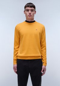 Geel gebreid trui met een ronde halslijn, geribbelde manchetten en boord. Klein logo op de borst. Gedirigeerd met zwarte broek.