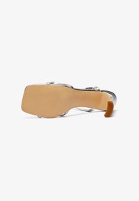Metallic silver high-heeled sandal with clear straps, block heel, and beige rubber sole. The sole has a textured finish for grip.