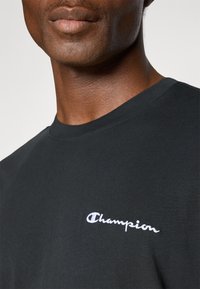 Schwarzes Baumwoll-T-Shirt mit Rundhalsausschnitt, ausgestattet mit einem weißen „Champion“-Logo-Stick auf der linken Seite. Glatte Textur und lässiges Design.