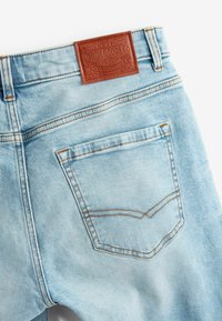 Jean en denim bleu clair avec une pièce en cuir brun texturé à l'arrière. Comprend des poches standard et un détail de couture contrastante jaune.