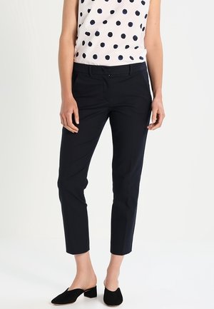 Femme portant un pantalon slim noir jusqu'à la cheville, un top blanc sans manches à pois noirs et des chaussures noires à talons bas à enfiler.