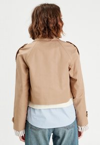Veste beige cintrée avec une finition lisse, dotée d'épaules épaulettes et d'un ourlet blanc contrastant, superposée sur une chemise à rayures bleues et blanches.