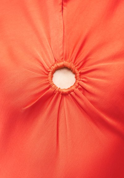 Κοντινό πλάνο πορτοκαλί υφάσματος σφιχτά συγκεντρωμένου γύρω από μια μικρή κυκλική άνοιγμα, που δείχνει υφή και λεπτομέρειες ραφής.