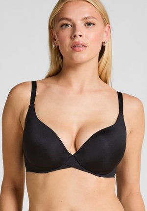 Blondynka nosząca czarny wyściełany biustonosz z cienkimi ramiączkami na jednolitym jasnoszarym tle.