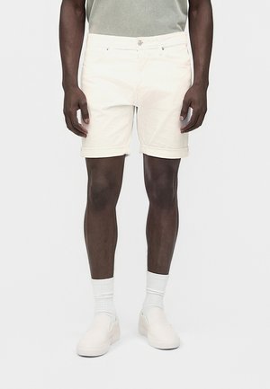 Mand iført lyse beige shorts med opsmøgede kanter, hvide sokker og hvide slip-on sko, står mod en ensfarvet lys baggrund.