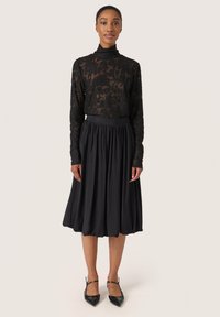 Černý krajkový top s dlouhým rukávem a vysokým výstřihem, doplněný volnou černou sukní ke kolenům. Outfit doplňují špičaté černé boty.