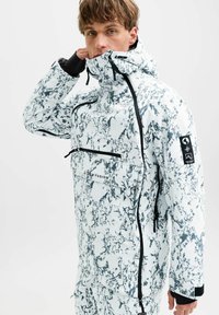 Jeune homme ajustant sa capuche, portant une veste d'hiver marbrée blanche et grise et un pantalon avec des détails de fermeture éclair noirs, sur un fond uni.