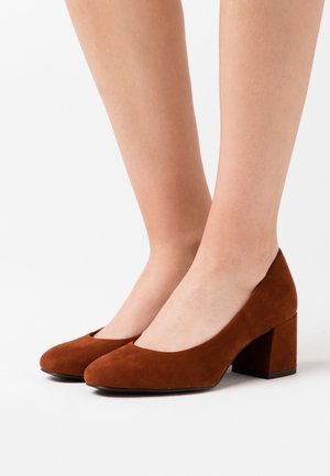 Escarpins en daim brun à talon carré avec un bout arrondi et une texture lisse. Dotés d'un talon de hauteur intermédiaire et d'un design minimaliste.