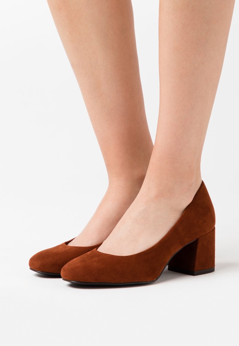 Escarpins en daim brun à talon carré avec un bout arrondi et une texture lisse. Dotés d'un talon de hauteur intermédiaire et d'un design minimaliste.